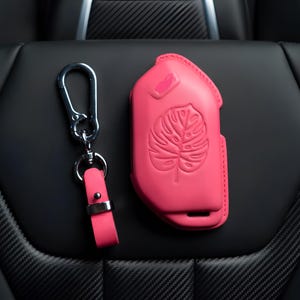 May include: A bright pink key fob cover and matching keychain set. The key fob cover features a raised monstera leaf design. The keychain has a pink leather strap and a silver-colored metal clasp.