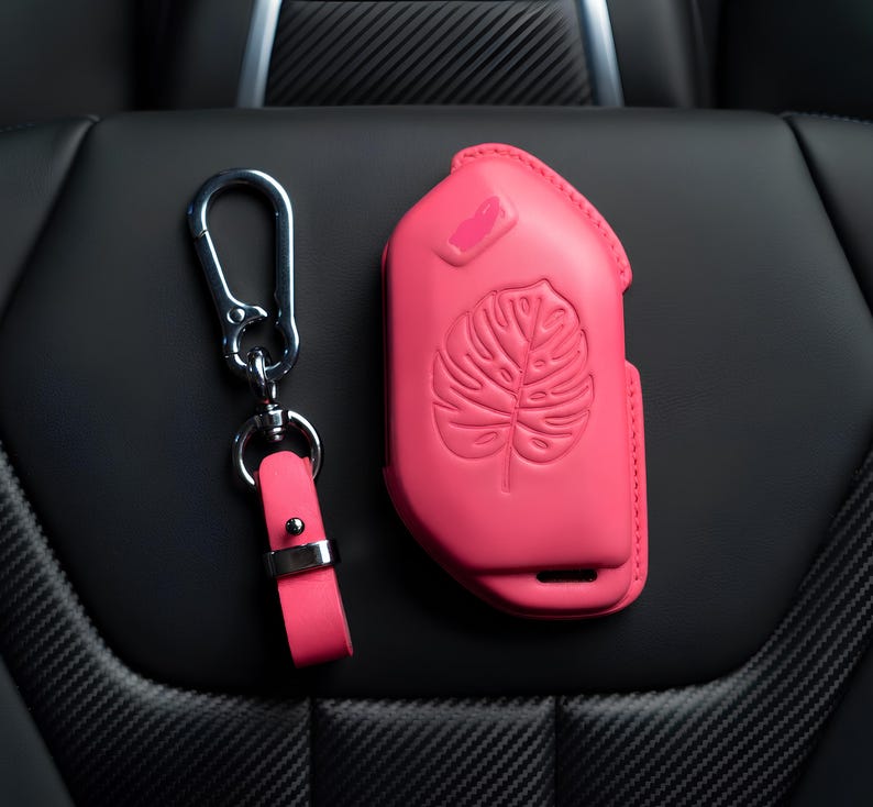 May include: A bright pink key fob cover and matching keychain set. The key fob cover features a raised monstera leaf design. The keychain has a pink leather strap and a silver-colored metal clasp.
