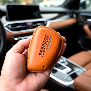 May include: Orange leather key fob cover with the text "ZR2 BISON" in black and silver. The cover is held in a hand, with a car interior in the background.