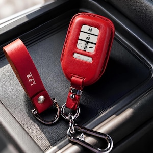 May include: A red leather key fob case and matching strap with silver hardware. The key fob case has white buttons with symbols and the word "HOLD". The strap is embossed with the initials "L.F.E."