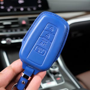 May include: A blue leather key fob cover with embossed buttons for lock, unlock, A/C, and other functions. The fob has a silver-colored metal ring and a matching blue strap. The background shows a car's interior.