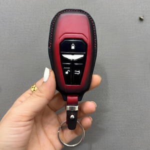 May include: A red and black leather car key fob cover with a key ring. The fob has a black panel with white symbols for lock, unlock, and other functions. The cover is stitched with red thread.
