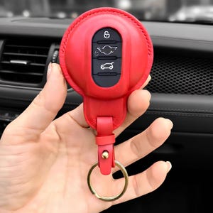 May include: A bright red leather key fob cover for a Mini Cooper car key. The cover has a circular design with a black panel featuring key symbols. A gold-colored key ring is attached to the fob.