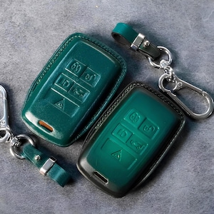 May include: Two teal leather key fob covers with embossed buttons and a key ring. One key fob cover has a silver carabiner clip. The other has a silver key ring. The key fobs are on a grey surface.