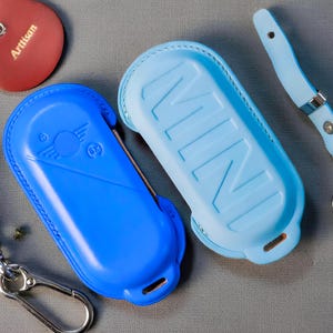 May include: Two blue leather key fob cases with silver carabiner clips. One is a solid bright blue, the other is a lighter blue with the word "MINI" embossed. A red cat-shaped leather tag with "Artisan" is also visible.