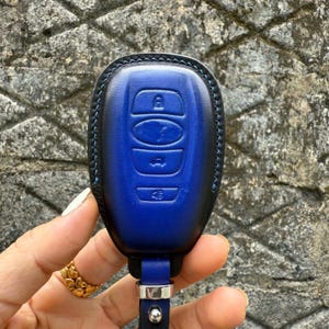 May include: A blue leather car key fob cover with a black border and blue stitching. The fob has embossed buttons for locking, unlocking, and other functions. A black leather strap with a silver-tone metal ring is attached.
