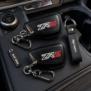 Puede incluir: Fundas de llavero y llaveros de cuero negro con herrajes plateados. Las fundas de llavero tienen un logotipo "ZR2" blanco y rojo. Un llavero dice "High Country" y otro "35AE Miata".
