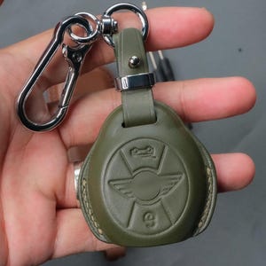 Peut inclure: Porte-clés en cuir vert olive avec mousqueton en métal argenté. Le porte-clés présente un logo en relief avec un design de voiture et d'ailes. Le porte-clés est conçu pour une clé de voiture.