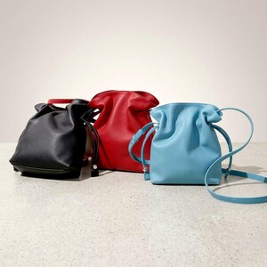 May include: Three leather drawstring bags in black, red, and light blue. The bags have a gathered top and a shoulder strap. The bags are sitting on a light gray surface.