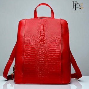 Peut inclure: Un sac à dos rouge vif avec une texture peau de crocodile. Le sac est doté d'une poignée supérieure, de bretelles réglables et d'une fermeture éclair. Le design comprend un motif central de crocodile en relief, ajoutant une touche unique.
