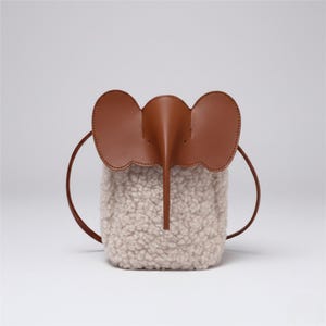 Puede incluir: Un bolso único y caprichoso con forma de elefante. El bolso presenta una cabeza de elefante de cuero marrón con orejas grandes y una trompa, sobre un cuerpo esponjoso de color crema. Tiene correas marrones.