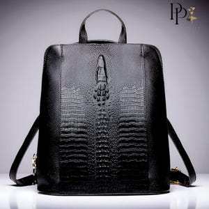 Puede incluir: Mochila de cuero negro con textura de piel de cocodrilo. La mochila tiene un asa superior, correas ajustables para los hombros y un diseño decorativo de cocodrilo en la parte delantera. La bolsa tiene forma rectangular y un diseño elegante y moderno.