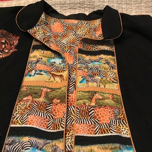 May include: Black garment with a collar and decorative panels featuring animal prints. The panels display giraffes, zebras, and leopard spots. A tiger face is embroidered on the left side. The garment has a tan trim.