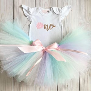 May include: A white onesie with a gold glitter 'one' and a pink seashell graphic. The onesie is paired with a rainbow tulle tutu with a pink satin bow.