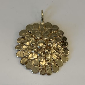Puede incluir: Un colgante dorado con un diseño floral. El colgante presenta una esfera central elevada rodeada de numerosos elementos en forma de pétalo, creando una apariencia texturizada y en capas. Tiene un pequeño lazo en la parte superior para una cadena.