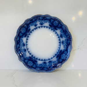 Può includere: Un piatto rotondo in ceramica con un centro bianco e un ampio bordo smerlato decorato con un motivo floreale blu intenso. Il bordo del piatto è rifinito con un sottile profilo dorato e il design complessivo presenta motivi fluidi e complessi.