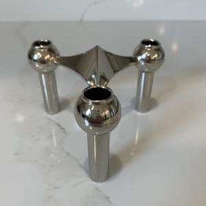 Peut inclure: Un bougeoir en métal argenté poli doté d'un design à trois pieds. Chaque pied est surmonté d'une coupelle à bougie sphérique, et les trois pieds se rejoignent en un point central, formant une structure triangulaire à la surface réfléchissante.