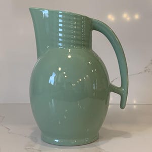 May include: A mint green ceramic pitcher with a rounded body and a curved handle. The pitcher has a spout for pouring and a textured band near the top. The pitcher is a decorative item for serving beverages.