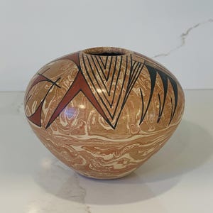 May include: A round, hand-painted ceramic vase with a marbled, tan and white base. The vase features geometric designs in black and red. The opening is a dark, solid color. This decorative piece is suitable for home decor.