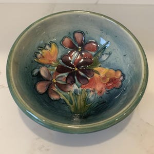 Könnte beinhalten: Eine Keramikschale mit handgemaltem Blumendesign. Die Schale hat eine teal-farbene Außenseite und eine hellblaue Innenseite. Das Blumendesign umfasst orange, rote und violette Blumen mit grünen Blättern. Die Schale ist rund und hat eine glänzende Oberfläche.
