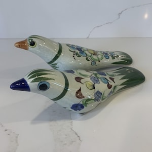 May include: Two ceramic bird ornaments. The birds are white with green, blue, and brown floral patterns. One bird has a blue beak, the other has an orange beak. The birds have black eyes and are positioned on a white surface.