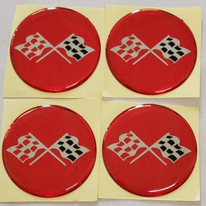May include: Four red, circular decals featuring a crossed checkered flag design. The flags are white and black, set against a vibrant red background. The decals are on a square, light-yellow backing.