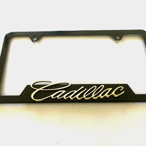 Puede incluir: Marco de matrícula de fibra de carbono negro con un logotipo dorado de Cadillac.