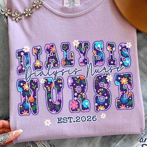 May include: A lilac Comfort Colors t-shirt with the words "DIALYSIS NURSE" in a floral design. The text "EST.2026" is below the main text. A brown hat and a floral necklace are also visible.