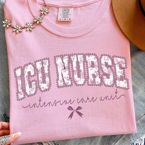 May include: A light pink t-shirt with the words "ICU NURSE" in a floral design. Below the text is "intensive care unit" in cursive. A small bow graphic is below the text. A floral necklace and a brown hat are also in the image.