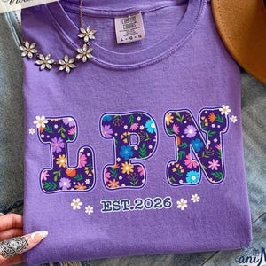 May include: A purple Comfort Colors t-shirt with the letters "LPN" in a floral design. The text "EST.2026" is below the letters. A floral necklace and a brown hat are also visible.