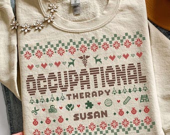 Puede incluir: Una sudadera de cuello redondo color arena con un patrón festivo navideño en rojo, verde y marrón. El diseño incluye el texto "OCCUPATIONAL THERAPY" y "SUSAN" junto a iconos de un caduceo, corazones, árboles y juguetes.