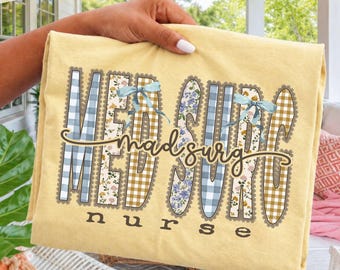 Puede incluir: Una camiseta de color amarillo pálido con el texto "med surg" en letras grandes y bloqueadas con un borde festoneado. Las letras alternan entre patrones de cuadros azules, flores y cuadros beige, acentuadas por dos lazos azul claro. La palabra "nurse" está impresa debajo.