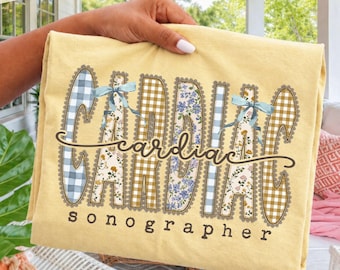 Puede incluir: Una camiseta de color amarillo pálido con la palabra CARDIAC en letras grandes con bordes festoneados, con patrones de cuadros azules, flores y cuadros marrones. Dos lazos azul claro están unidos a las letras. La palabra sonographer está debajo.