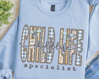 Puede incluir: Una sudadera de cuello redondo en color azul claro con el texto "child life specialist" en una fuente marrón oscura. Las letras están rellenas con patrones de cuadros vichy azules, cuadros amarillos y flores con lazos.