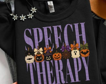 Puede incluir: Una sudadera negra de cuello redondo con el texto SPEECH THERAPY en cuadros morados. Debajo del texto hay siete manzanas de caramelo decoradas con lazos, incluyendo diseños de momia, gato negro, sombrero de bruja, calabaza, telaraña, fantasma y murciélago.