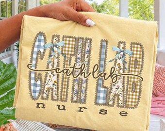 Puede incluir: Una camiseta de color amarillo pálido con el texto "cath lab nurse" en una fuente cursiva de color marrón oscuro. Las letras están rellenas con patrones de cuadros azules, cuadros amarillos y flores, con dos lazos azul claro.