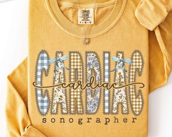 Puede incluir: Una camiseta de manga larga color amarillo mostaza con el texto "cardiac sonographer" en letra cursiva y de imprenta marrón. Las letras tienen rellenos de cuadros vichy azules, flores y cuadros beige, con lazos azul claro.