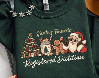 Puede incluir: Una sudadera de cuello redondo color verde bosque con un estampado gráfico que incluye el texto Santa's Favorite Registered Dietitian. El diseño muestra un árbol de Navidad decorado, una taza de chocolate caliente, un hombre de jengibre y a Santa.