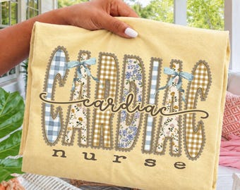 Puede incluir: Una camiseta de color amarillo pálido con el texto "cardiac nurse" en letras de color marrón oscuro. La palabra "cardiac" aparece en letras grandes y decorativas con patrones alternos de cuadros azules, flores y cuadros beige, acentuada por dos pequeños lazos azul claro.
