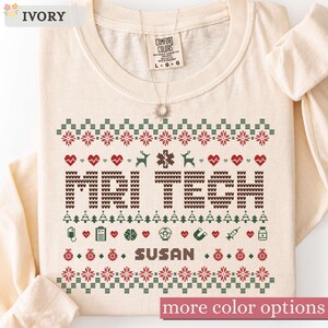 May include: An ivory long-sleeve shirt featuring a festive knit-style graphic with the text MRI TECH and SUSAN. The design includes red hearts, green trees, medical icons like a brain and syringe, and geometric patterns in red, green, and brown.