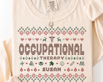 Puede incluir: Una camiseta de manga larga color marfil con un gráfico estilo tejido festivo y el texto OCCUPATIONAL THERAPY SUSAN. El diseño incluye patrones rojos y verdes de corazones, renos, árboles, adornos e iconos de terapia.