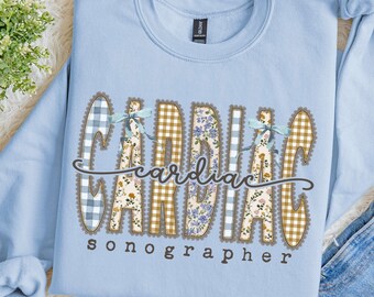 Puede incluir: Una sudadera de cuello redondo en color azul claro con el texto Cardiac Sonographer en una fuente decorativa. Las letras están rellenas con varios patrones, incluyendo cuadros vichy azules, cuadros amarillos y estampados florales.