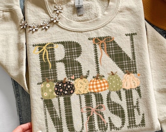 Puede incluir: Una sudadera de cuello redondo color arena con el texto RN NURSE en un patrón de cuadros verde oscuro. Las letras están decoradas con varias calabazas ilustradas en tonos naranja, verde y crema, acentuadas con pequeños lazos amarillos y rosas.
