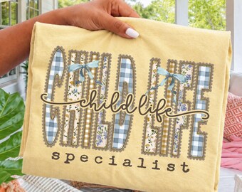 Puede incluir: Una camiseta de color amarillo pálido con el texto "child life specialist" en letra cursiva y de imprenta marrón oscuro. Las letras tienen rellenos de cuadros vichy azules y blancos, amarillos y blancos, y flores con lazos azules.