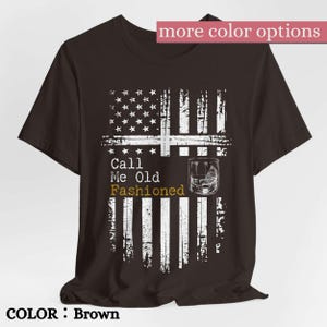 May include: A brown short-sleeve t-shirt featuring a distressed white graphic of an American flag. The design includes a cocktail glass illustration and the text Call Me Old Fashioned, with the word Fashioned printed in a gold-toned yellow color.