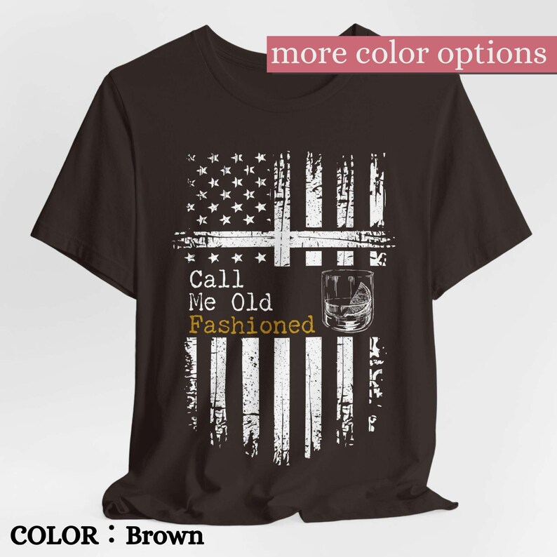 May include: A brown short-sleeve t-shirt featuring a distressed white graphic of an American flag. The design includes a cocktail glass illustration and the text Call Me Old Fashioned, with the word Fashioned printed in a gold-toned yellow color.