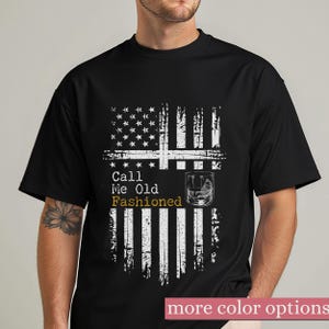 May include: A black short-sleeved t-shirt featuring a distressed white graphic of the American flag. The design includes the text "Call Me Old Fashioned" in white and gold lettering, accompanied by an illustration of a glass of whiskey.
