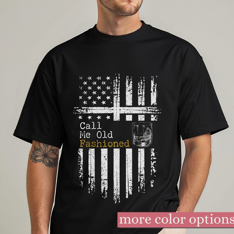 May include: A black short-sleeved t-shirt featuring a distressed white graphic of the American flag. The design includes the text "Call Me Old Fashioned" in white and gold lettering, accompanied by an illustration of a glass of whiskey.