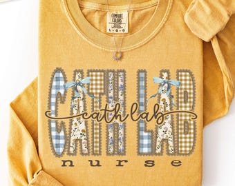 Puede incluir: Una camiseta de manga larga color amarillo mostaza con una etiqueta Comfort Colors en el cuello. El frente presenta el texto "cath lab" en una fuente cursiva marrón, con las letras "CATH LAB" rellenas de patrones florales y de cuadros azules.
