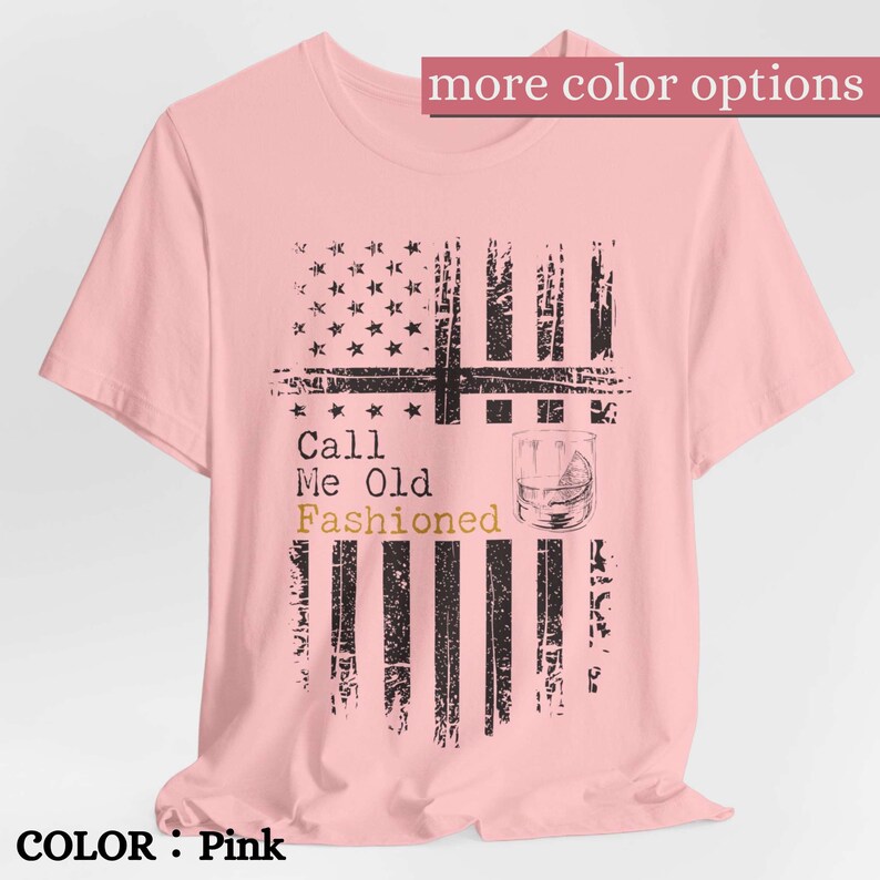 May include: A light pink short-sleeve t-shirt featuring a distressed black graphic of an American flag. The design includes the text "Call Me Old Fashioned" with the word "Fashioned" in gold, alongside a line drawing of a cocktail glass.